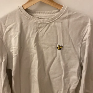 Vit långärmad tröja från Lyle & Scott - Säljer en stilren vit långärmad tröja från Lyle & Scott med deras ikoniska logga broderad på bröstet. Tröjan är tillverkad i ekologisk bomull och har en ribbad krage. Perfekt för en avslappnad och snygg look.