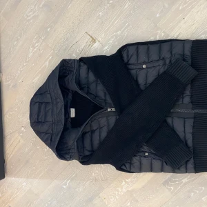 Svart dunjacka från Moncler - Snygg svart dunjacka från Moncler med huva och dragkedja. Jackan har långa ärmar i ribbat material och två fickor med knappar framtill. Perfekt för kyligare dagar.