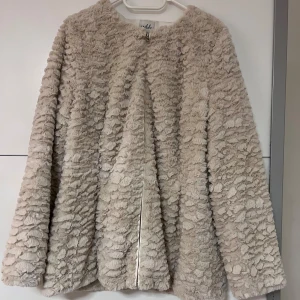 Beige teddyjacka från Isolde - Mysig beige teddyjacka från Isolde med dragkedja framtill. Jackan har en fluffig textur och långa ärmar, perfekt för kyligare dagar. Den är fodrad med ett glansigt material som ger en lyxig känsla. Ett måste för garderoben! 