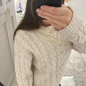 Beige stickad tröja - Mysig beige stickad tröja med kabelstickat mönster. Perfekt för kyliga dagar med sin varma och bekväma design. Tröjan har långa ärmar och en rund halsringning.