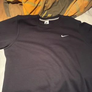 Svart tröja från Nike - Säljer en stilren svart tröja från Nike med deras ikoniska logga på bröstet. Tröjan har en klassisk passform och är perfekt för en avslappnad look. Tillverkad i ett mjukt material för extra komfort.