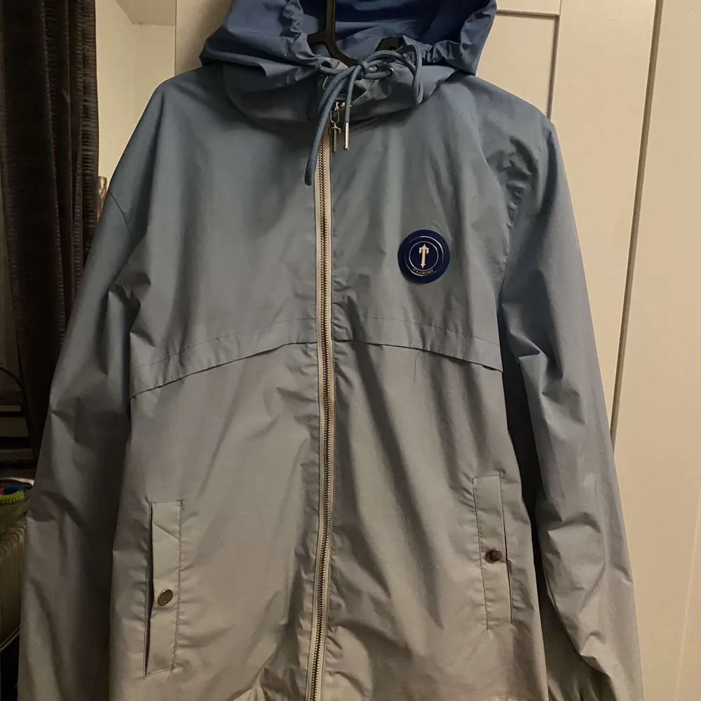 Säljer en blå jacka från Trapstar med huva och dragkedja. Jackan har ett tryck på ryggen och en logotyp på bröstet. Perfekt för en stilren look. Passar även storleken M . Takit.