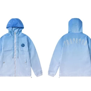 Blå vindjacka med broderad text - Snygg ljusblå vindjacka med huva och dragkedja framtill. Jackan har en broderad text på ryggen och en rund logotyp på bröstet. Perfekt för blåsiga dagar.