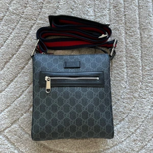 Grå axelväska från Gucci - Snygg grå axelväska från Gucci med det klassiska GG-mönstret. Väskan har en justerbar axelrem i rött och blått samt en praktisk dragkedja framtill. Perfekt för att bära med sig det viktigaste med stil. HELT NY och Oanvänd. Perfekt för resor och sommaren!