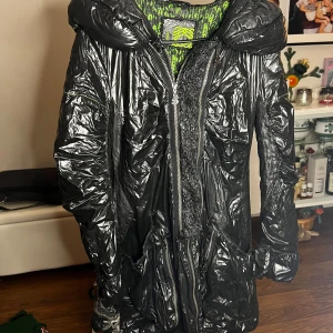 Svart glansig jacka från Portalm - Snygg svart glansig jacka från Portalm med en cool grönmönstrad insida. Jackan har en stor huva och praktiska fickor. Perfekt för att sticka ut i mängden med sin unika stil. Den har dragkedja framtill för enkel stängning.