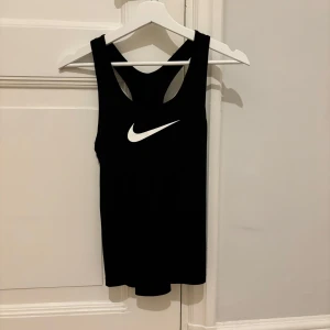 Svart träningslinne från Nike - Säljer en svart topp från Nike med Dri-FIT-teknologi. Toppen har en klassisk design med ett vitt Nike-logotyptryck på framsidan. Perfekt för träning med sin bekväma passform och fukttransporterande material.