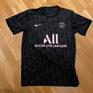 Svart PSGxDIOR fotbollströja - Snygg svart PSG fotbollströja från Nike med rosa detaljer och klubbmärke. Tröjan har ett diskret mönster och korta ärmar. Perfekt för alla PSG-fans som vill visa sitt stöd med stil. Storleken är M men kan passa S.                                                  Pris kan sänkas vid snabb affär.                                         TAR EMOT BYTEN