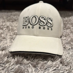 Beige keps från Hugo Boss - Snygg beige keps från Hugo Boss med broderad logga framtill. Kepsen har en justerbar rem baktill med metallspänne för perfekt passform. Perfekt för en stilren look.