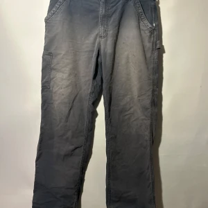 Grå arbetsbyxor från Carhartt - Ett par Carhartt byxor i modellen Original Dungaree fit. Byxorna har fickor på sidorna och bakfickor. Byxorna är mycket slitna med hål på båda sidor, även solblekta.