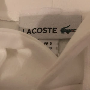 Vit hoodie från Lacoste - Säljer en stilren vit hoodie från Lacoste med klassisk krokodillogga på bröstet. Tröjan har en rymlig ficka framtill och justerbar huva med snören. Perfekt för en avslappnad stil.