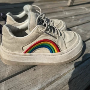Beige sneakers med regnbågsbroderi - Snygga beige sneakers från Eytys med ett färgglatt regnbågsbroderi på sidan. Skorna har snörning och en bekväm, tjock sula. Perfekta för en avslappnad stil med en touch av färg.