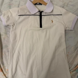 Vit pikétröja från Ralph Lauren - Säljer en stilren vit pikétröja från Ralph Lauren med dragkedja och broderad logga på bröstet. Tröjan har korta ärmar och en klassisk krage med kontrastfärgade detaljer. Perfekt för en avslappnad men ändå elegant look.