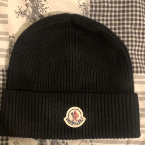 Moncler mössa - Snygg svart ribbad mössa från Moncler med deras ikoniska logotyp framtill. Perfekt för att hålla värmen under kyliga dagar. Stilren design som passar till det mesta.