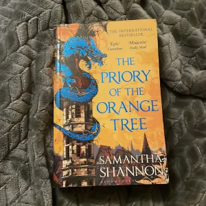 The Priory of the Orange Tree - Säljer boken 'The Priory of the Orange Tree' av Samantha Shannon. Denna internationella bästsäljare är en episk fantasyroman med en vacker omslagsdesign i orange och blått, prydd med en drake. Perfekt för fantasyälskare som söker en storslagen berättelse. Lite sliten och har några defekter. 