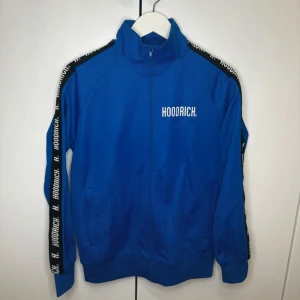 Blå träningsjacka från Hoodrich - Snygg blå träningsjacka från Hoodrich med dragkedja och hög krage. Jackan har svarta band med vit text längs ärmarna och en logga på bröstet. Perfekt för en sportig look!