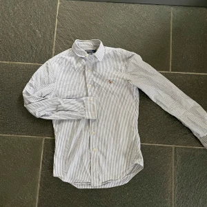 Randig skjorta från Ralph Lauren - Snygg vit och blå randig skjorta från Ralph Lauren med klassisk krage och knappar framtill. Skjortan har långa ärmar och en liten broderad logga på bröstet. Perfekt för en stilren look!