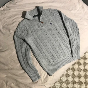 Ralph Lauren kabelstickad tröja - Säljer en stilren grå stickad tröja från Ralph Lauren med kabelstickat mönster och en liten broderad logga på bröstet. Tröjan har en dragkedja vid halsen och ribbade muddar. Perfekt för kyliga dagar! 💯top kvalitet som vanligt 💯