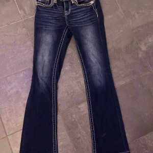 Blå bootcut jeans från Miss Me - Snygga blå bootcut jeans från Miss Me med dekorativa bakfickor och kontrastsömmar. Perfekta för en avslappnad stil med en touch av detaljer. Midjan är låg och passformen är normal.