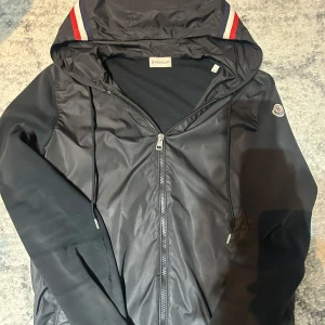 Svart jacka från Moncler - Säljer en snygg svart jacka från Moncler med huva och dragkedja. Jackan har Moncler-logga på ärmen och detaljer i rött och vitt på huvan. Perfekt för en stilren look.