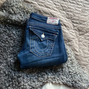 Blå jeans från True Religion - Snygga blå jeans från True Religion med klassisk femficksdesign och dekorativa sömmar på bakfickorna. Tillverkade i USA, dessa jeans har en lågmidjad passform och en bootcut-stil. Mått i midjan är 88 cm och insida lår är 65 cm