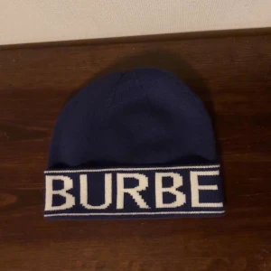 Mörkblå mössa från Burberry - Snygg mörkblå mössa från Burberry med vit textdetalj. Perfekt för att hålla värmen med stil. Den har en klassisk passform och är tillverkad i ett mjukt material.