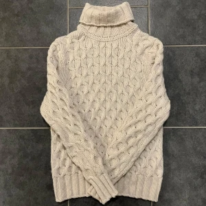 Beige stickad polotröja från Morris - Säljer en mysig beige stickad polotröja från Morris. Tröjan har ett klassiskt flätmönster och en hög polokrage som håller dig varm under kyliga dagar. Perfekt för höst och vinter!