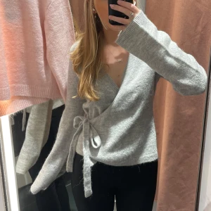 Grå omlottkofta med knytband - Säljer en mysig grå omlottkofta med knytband i midjan. Koftan har långa ärmar och är perfekt för kyligare dagar. Den är både stilren och bekväm, vilket gör den till ett mångsidigt plagg i garderoben. Använd 2 gånger