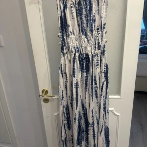 Vit och blå långklänning - Snygg långklänning i vitt och blått med ett unikt mönster. Klänningen har en smockad midja för en bekväm passform och är ärmlös, vilket gör den perfekt för varmare dagar. Materialet är lätt och luftigt.