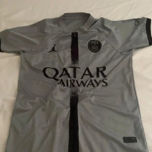 Grå PSG fotbollströja från Jordan - Snygg grå PSG fotbollströja med svart vertikal rand och Qatar Airways-logga på framsidan. Tröjan har korta ärmar med svarta detaljer och är tillverkad i ett lätt och bekvämt material. Perfekt för fotbollsfans! 7/10 skick. Anände den ganska lagom i sommaren