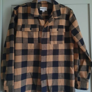 Rutig flanellskjorta - Snygg rutig flanellskjorta i senapsgul och svart. Skjortan har långa ärmar och knappar framtill samt två bröstfickor med knappar. 