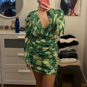 Grön blommig klänning - Säljer en snygg grön klänning med blommigt mönster från Adoore. Den är kort med rynkade detaljer på sidan. Klänningen är aldrig använd så i helt nytt skick! 