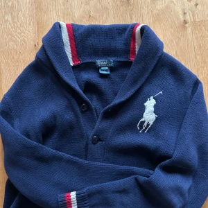 Mörkblå kofta från Ralph Lauren - Säljer en snygg mörkblå kofta från Ralph Lauren med det ikoniska vita polospelarlogotypen på bröstet. Koftan har en krage med röd och vit randig detalj samt knappar framtill. Perfekt för en stilren look! Skulle säga att den passar någon Melker 170-175👊👊 skriv vid funderingar