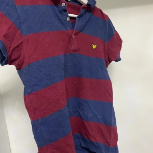 Randig pikétröja från Lyle & Scott - Säljer en randig pikétröja från Lyle & Scott i blått och vinrött. Tröjan har korta ärmar och en klassisk krage med knappar. Perfekt för en avslappnad stil.