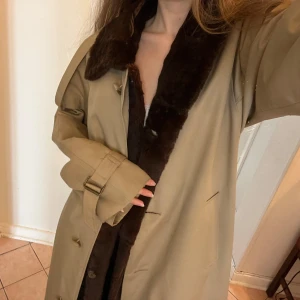 Beige trenchcoat med pälsdetaljer - Snygg beige trenchcoat med mörkbrun innerpäls. Jackan har långa ärmar och ett bälte i midjan för en justerbar passform. En stilren design med knappar, i väldigt bra skick. Några små defekter är mindre fläckar på ärmen och nån knapp som sitter lite löst 🌸 Kontakta vid frågor 💌(pris kan diskuteras)