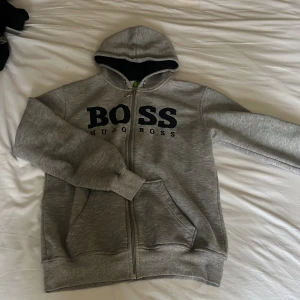 Grå hoodie från Hugo Boss - Säljer en stilren grå hoodie från Hugo Boss med dragkedja och stor logga på bröstet. Tröjan har en bekväm passform med långa ärmar och en praktisk huva. Perfekt för en avslappnad look. Pris diskuteras gärna