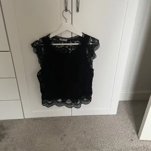 Svart spetsblus - Säljer en elegant svart spetsblus med vackra blommönster. Blusen är ärmlös och har fina spetsdetaljer runt halsen och ärmsluten. Perfekt för en stilfull look.