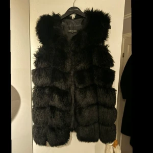 Svart pälsväst från Verdallo - Säljer en lyxig svart pälsväst från Verdallo. Västen har en fluffig och elegant design med en huva som ger extra värme och stil. Perfekt för att hålla sig varm och trendig under kallare dagar.