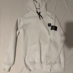 Vit stone Island hoddie - Säljer en stilren vit hoodie från Stone Island med deras ikoniska logotyp på ärmen. Hoodien har en praktisk känguruficka och justerbar huva med svarta snören. Pris kan diskuteras