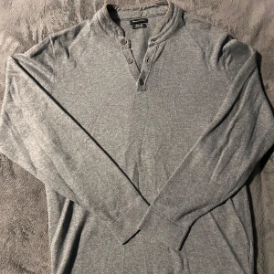 Grå tröja från Massimo Dutti - Säljer en stilren grå tröja från Massimo Dutti med knappar vid halsen. Tröjan har långa ärmar och en mjuk krage med en fin detalj i form av en kontrasterande kant. Perfekt för en avslappnad men ändå elegant look.