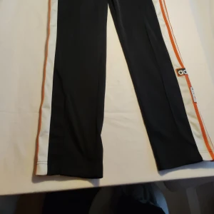 Svarta träningsbyxor från Adidas - Snygga svarta träningsbyxor från Adidas med vita och orange ränder längs sidorna. Byxorna har knappar längs benen och elastisk midja för extra komfort. Perfekta för en sportig look.