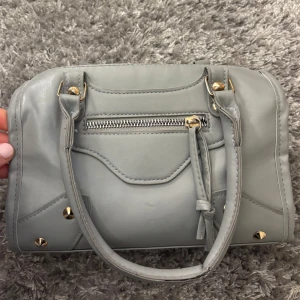 Grå handväska med dragkedja och nitar - Snygg handväska med nitar i guld. Väskan har två handtag och ett långt band men det har släppt på ena sidan men går nog enkelt att laga💕
