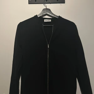 Svart Tröja, lite som en tunn overshirt. - Snygg svart jacka overshirt med dragkedja framtill. Tröjan  har en minimalistisk design och långa ärmar, perfekt för en stilren look. Passar bra till både vardag och mer uppklädda tillfällen.