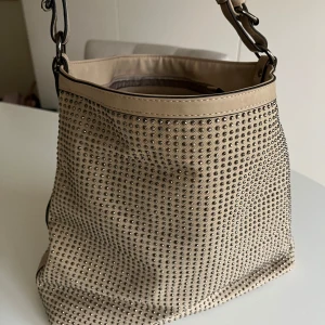 Beige axelväska med nitar - Snygg beige axelväska dekorerad med nitar. Väskan har en avtagbar axelrem och flera fack med dragkedja inuti för enkel organisering. Perfekt för att ge din outfit en edgy touch. Nyskick. Använd fåtal ggr!