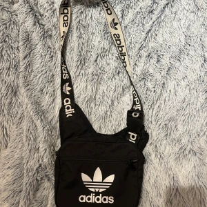 Svart axelväska från Adidas - Snygg svart axelväska från Adidas med det klassiska trefoil-loggan i vitt. Väskan har en justerbar axelrem med Adidas-tryck och en praktisk dragkedja framtill. 3 fack och väldigt rymlig. Perfekt för att bära med sig det nödvändigaste på språng.