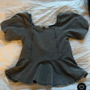 Grå topp med puffärmar - Säljer en snygg grå topp med puffiga kortärmar och volangdetalj i nederkant. Toppen har en fyrkantig halsringning och är tillverkad i ett jeansliknande material. Perfekt för en trendig look!