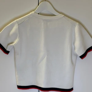 Vit kortärmad topp med svarta och röda detaljer - Snygg vit topp med korta ärmar och stilrena svarta och röda ränder vid ärmslut och nederkant. Perfekt för en trendig och avslappnad look. Från zara. 