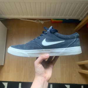 Blå sneakers från Nike - Säljer ett par blå sneakers från Nike med vit sula och vit swoosh-logga. Skorna har snörning och är tillverkade i mocka. Perfekta för en avslappnad stil.
