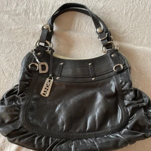 Svart axelväska från DKNY - Snygg svart axelväska från DKNY i skinn med silverdetaljer och en praktisk dragkedja. Väskan har en stilren design med dekorativa sömmar och en rymlig insida med flera fack. Perfekt för att bära med sig det viktigaste med stil. Något missfärgad (rosa ) på ena sidan invändig, men inget som påverkar. 