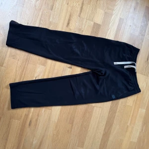 Svarta byxor från Acne Studios - Snygga svarta byxor från Acne Studios med elastisk midja och justerbar snörning i rött och vitt. Byxorna har en praktisk bakficka med knappdetalj. Perfekta för en avslappnad stil. Mjukis byxor, storlek XS, passar S.