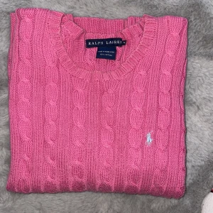 Rosa stickad tröja från Ralph Lauren - Säljer en snygg rosa stickad tröja från Ralph Lauren i 100% bomull. Tröjan har ett klassiskt kabelstickat mönster och den ikoniska loggan broderad på bröstet. Perfekt för en stilren look! Även perfekt som alla hjärtans dag present😉